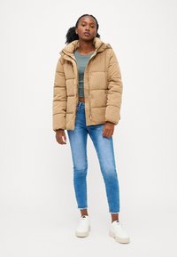 Bežna puff jakna s kapuco, z dikiranim vzorcem in zadrgo, oblečena čez zeleno majico in kombinirana z modrimi kavbojkami ter belimi športnimi copati.