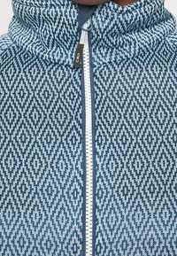 Blue patterned zip-up jacket featuring a diamond texture design, contrasting zipper, and a high collar. Made from soft, textured fabric.