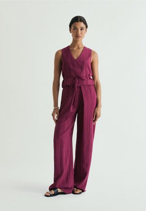 Donna in piedi con un gilet smanicato rosa intenso a righe sottili con cintura in vita e pantaloni ampi abbinati, indossa sandali neri.