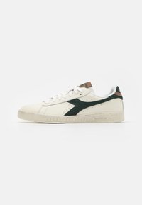 Hvit lær sneaker med grønn semsket akcent, teksturert gummisåle og brun hældetalj. Snøring med perforeringer på siden.