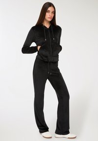 Młoda kobieta z długimi brązowymi włosami, w czarnej welurowej bluzie na zamek i dopasowanych spodniach, w białych sneakersach, stojąca z rękami w kieszeniach.