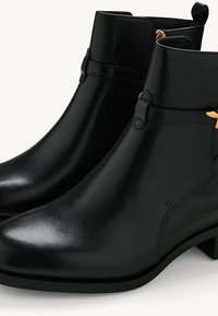 Bottes courtes en cuir noir avec talons bas, fermeture éclair latérale et détail de boucle dorée près du bord supérieur.