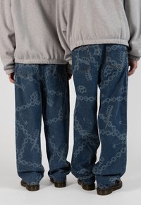 Blue denim jeans with a loose fit feature a chain and hardware pattern in a lighter shade. Back pockets visible, worn with gray sweatshirts.