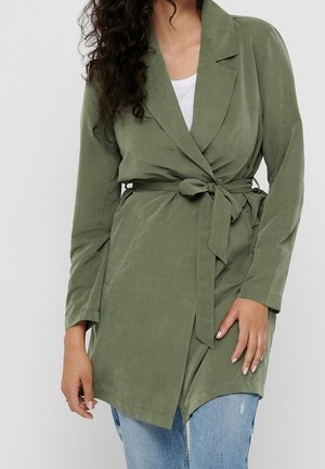 Donna che indossa un cappotto avvolgente verde oliva con cintura sopra una maglia bianca e jeans chiari strappati, in piedi davanti a uno sfondo semplice.