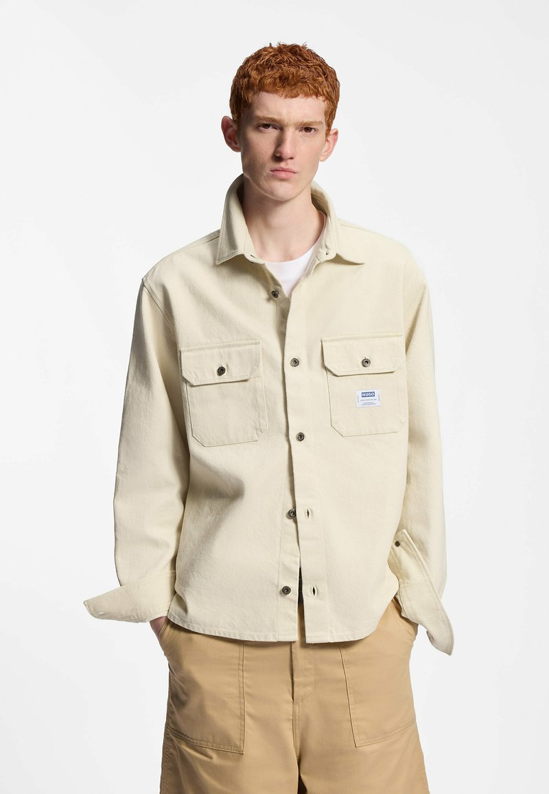 Jeune homme aux cheveux roux portant une chemise beige à boutons avec poches sur la poitrine et un pantalon cargo beige, debout les mains dans les poches.
