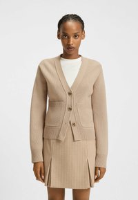 Beżowy kardigan z teksturowanym dzianiną, dekoltem w kszta�łcie litery V, dwoma przednimi kieszeniami i trzema guzikami. W zestawie z pasującą spódnicą w paski.