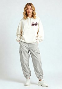 Felpa con cappuccio color crema con logo "Amour", abbinata a pantaloni cargo grigio chiaro con tasche laterali e polsini elasticizzati. Sneakers bianche completano l'outfit.