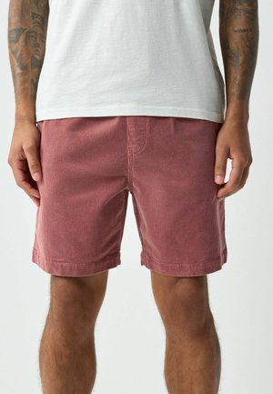 Mann trägt staubrosafarbene Cord-Shorts und ein weißes T-Shirt, zeigt tätowierte Unterarme und entspannt positionierte Hände.