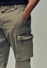 Cargo hlače u svijetloj khaki boji, izrađene od glatke tkanine. Dizajnirane s bočnim džepom s patentnim zatvaračem i opuštenim krojem.