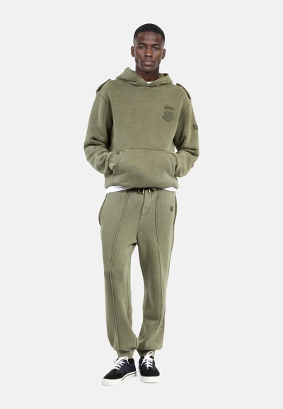 Felpa con cappuccio verde oliva e pantaloni jogger coordinati, con una texture morbida, tasca a marsupio e dettagli di cucitura. Abbinati a sneakers nere.