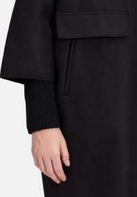 Czarny wełniany płaszcz o pudełkowym fasonie, z dużymi rękawami i dzianinowym wykończeniem mankietów. Posiada kieszeń naszywaną z przodu.