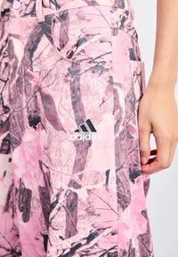 Rózsaszín álcázó nadrág, amely világos és sötét árnyalatok keverékéből készült, az oldalán fehér Adidas logóval és praktikus zsebekkel.