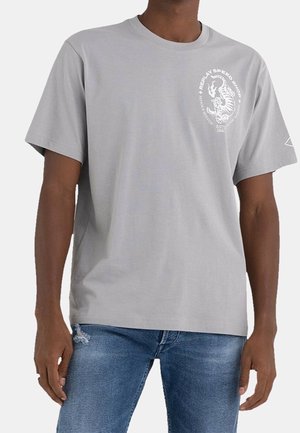 Mies, który nosi jasnoszary T-shirt z białym nadrukiem tygrysa i tekstem na piersi, w połączeniu z niebieskimi dżinsami.