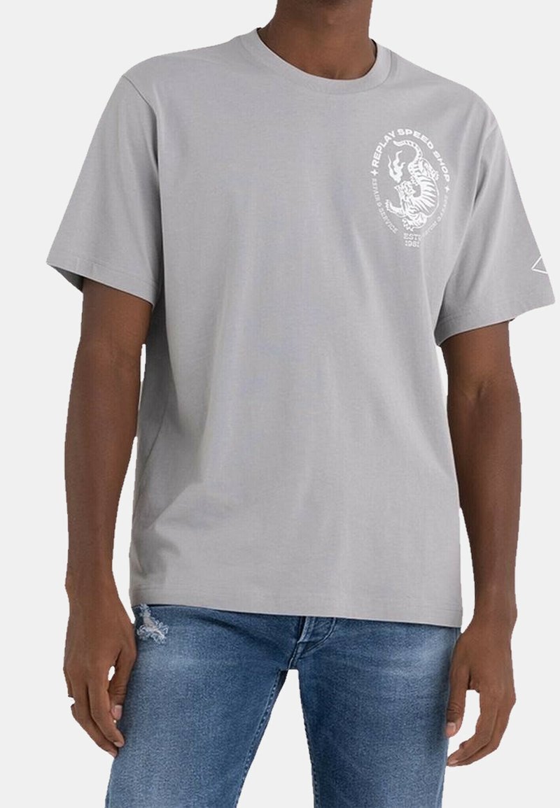 Hombre vestido con una camiseta gris claro que lleva una gráfica de un tigre blanco y texto en el pecho, combinada con jeans azules.