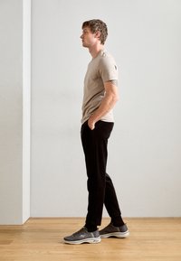 Camiseta ajustada de color beige claro, pantalones de pana negros, zapatillas deportivas grises. Perfil de pie contra una pared lisa, con las manos en los bolsillos.