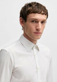 Camicia bianca con colletto classico a punta, texture liscia e chiusura a bottoni. Presenta maniche lunghe con polsini abbottonati.
