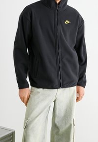 Man som bär en svart Nike zip-up fleecejacka med gult logotyp, i kombination med ljusgrå cargo byxor mot en neutral bakgrund.