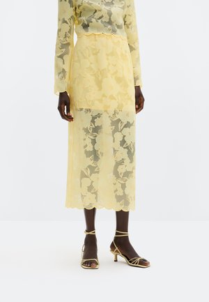 Modell iført gul, gjennomsiktig blomstrete midiskjørt med matchende topp og sandaler med lave hæler og stropper foran hvit bakgrunn.