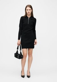 Γυναίκα που φοράει μαύρο μίνι φόρεμα με μακριά μανίκια και φερμουάρ στο μπροστινό μέρος, κρατώντας μαύρη τσάντα με τρουκς και φορώντας μαύρες μυτερές γόβες.