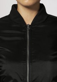 Giacca bomber nera con una texture liscia e lucida, colletto rialzato e chiusura frontale con zip. Design minimalista senza decorazioni aggiuntive.