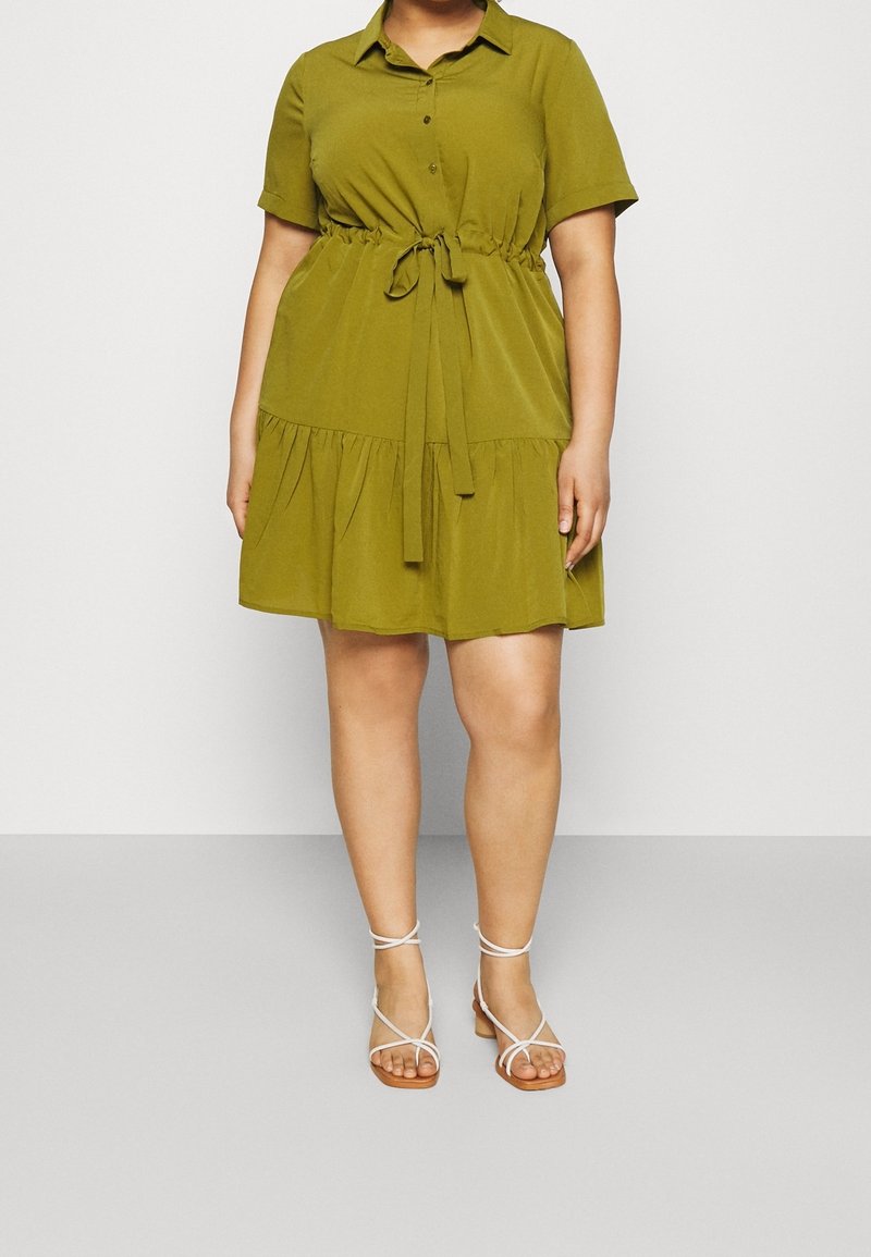 Femme portant une robe verte olive à manches courtes avec une ceinture à nouer et des sandales blanches à brides, debout devant un fond clair uni.