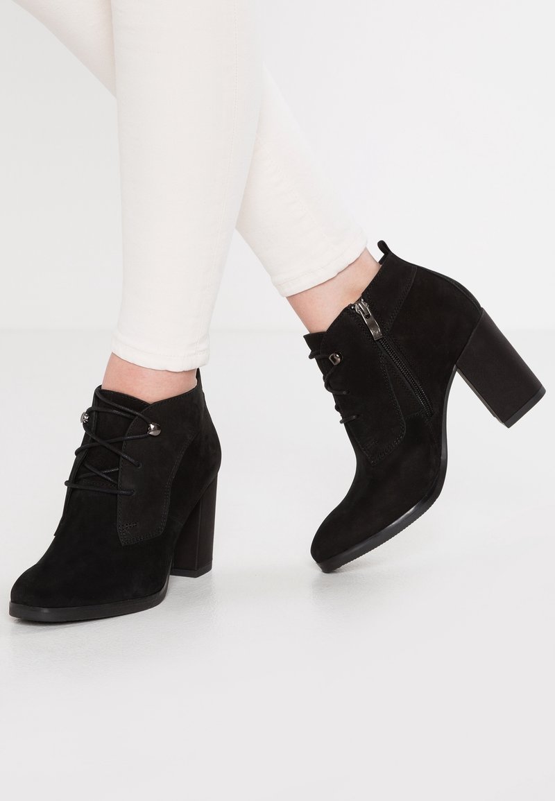 Bottes cheville en daim noir avec talons épais, fermetures éclair latérales et lacets, portées avec un pantalon blanc ajusté.