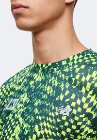 Κοντινό πλάνο ενός ατόμου που φοράει αθλητική μπλούζα σε νέον πράσινο και μαύρο μοτίβο με λογότυπα New Balance σε ανοιχτό φόντο.