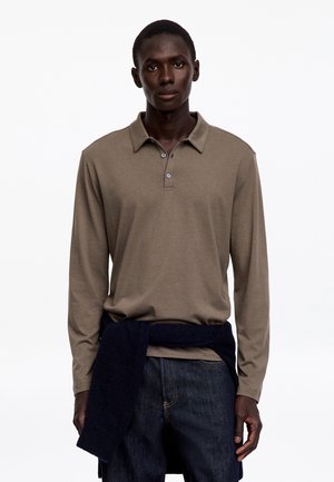 Homme portant un polo marron à manches longues et un jean foncé avec un pull noir noué autour de la taille, debout devant un fond blanc.