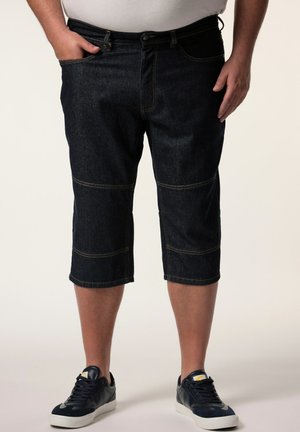 Mand iført mørkeblå knælange denimshorts og sorte sneakers, der står med den ene hånd i lommen og den anden ved siden.