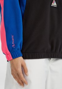 Fleecejakke med lyseblå og lyserøde ærmer, sort krop, elastiske manchetter og Polartec-branding. Trefarvet logo på brystet.