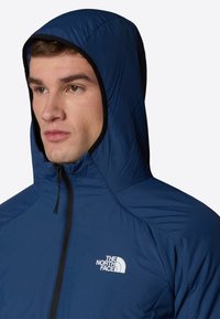 Marinblå huvjacka i ett mjukt och lätt material. Har en framficka med dragkedja och en vit logotyp på vänster bröst.