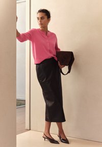 Lyserød cardigan med knapper, sort lædernederdel i midi-længde og sorte slingback-hæle. Krøllet hår og holder en brun taske mod en neutral baggrund.