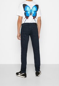 Vit t-shirt med en stor blå fjärilsgrafik på baksidan, kombinerad med mörkblå jeans och svarta gymnastikskor med vita detaljer.