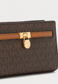 Brown patterned wallet made of textured material. Features a gold-tone clasp with a lock and tan leather strap. Logo embossed on the side.