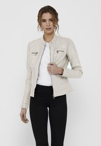 Giacca in pelle beige con tasche a zip sul petto, design aderente e colletto alla coreana. Indossata sopra una camicia bianca e jeans neri.