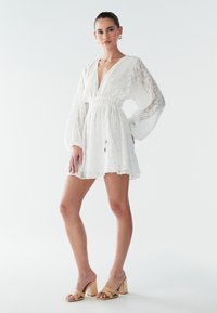 Vit kort klänning med djup V-ringning, långa puffärmar och dragsko i midjan. Har texturerat tyg och lager på fållen. Bärs med bruna sandaler med klossklack.