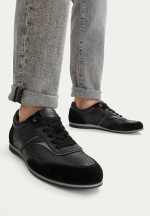 Schwarze, perforierte Ledersneaker, getragen mit hochgekrempelten grauen Jeans aus Denim vor weißem Hintergrund.