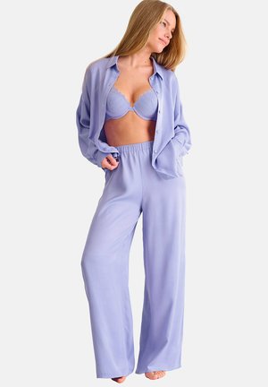 Femme portant un soutien-gorge lilas, un pantalon large assorti et une chemise à manches longues déboutonnée, posant pieds nus sur un fond blanc.