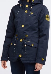 Marinblå vattentät jacka med huva, gula knappar, dragsko i midjan med träpärlor och en broderad logotypapplikation på ärmen.