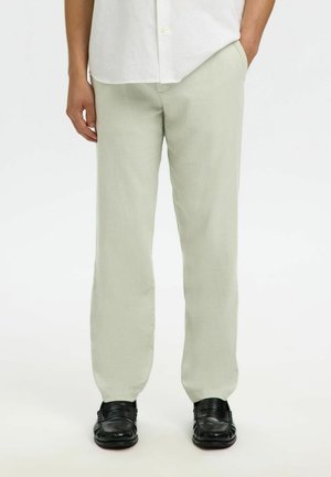 Homme portant un pantalon beige clair coupe droite, chemise blanche, mocassins en cuir noir, debout avec une main dans la poche sur fond blanc.