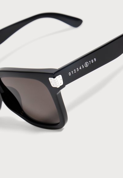Lunettes de soleil à monture noire avec verres foncés et motif numérique blanc de 0 à 9 sur la branche, dotées d’un détail de charnière argentée.
