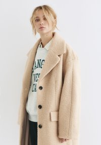 Beżowy płaszcz wełniany z wiskozą z wyciętym kołnierzem, dużymi czarnymi guzikami i bocznymi kieszeniami, noszony na szarym swetrze z nadrukowanym tekstem.