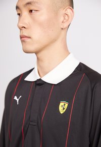 Juodas polo marškinėliai su raudonomis juostelėmis, balta apykakle, su Puma logotipu ir Ferrari emblemą ant krūtinės. Tekstūruotas audinio dizainas.