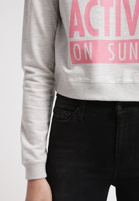 Šedá zkrácená mikina s velkým růžovým nápisem "ACTIVE ON SUN" na přední straně, v kombinaci s černými džíny s vysokým pasem.