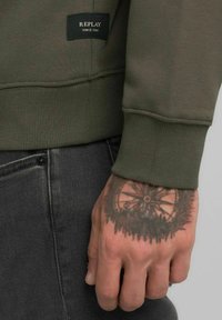Felpa verde oliva con polsini e orlo a coste, con etichetta nera "REPLAY". Jeans in denim nero visibili con dettaglio a bottone e un tatuaggio sulla mano.