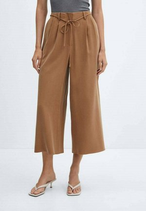 Woman wearing high-waist, wide-leg camel pants with a tied belt and off-white heeled sandals on a white background.