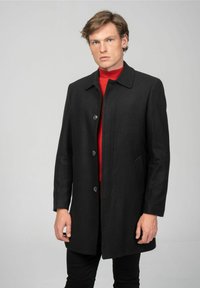 Zwarte wollen jas met een rechte snit, kraag en vijfknops sluiting. Draagt over een rode coltrui, heeft zijzakken en een gladde textuur.