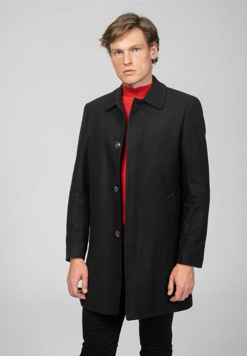 Zwarte wollen jas met een rechte snit, kraag en vijfknops sluiting. Draagt over een rode coltrui, heeft zijzakken en een gladde textuur.