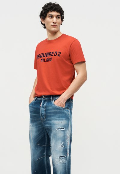 Νεαρός άνδρας στέκεται με τα χέρια στις τσέπες, φοράει πορτοκαλί μπλουζάκι "DSQUARED2 MILANO" και φθαρμένο μπλε τζιν μπροστά σε απλή φόντο.