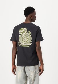 Black cotton T-shirt featuring a green graphic design of a skeleton and tools on the back. Short sleeves and crew neckline. Casual fit.